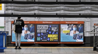 A photo of a Philly subway station. A woman is standing in front a three sheet poster display. The poster in the middle is advertising BlackStar Film Festival.