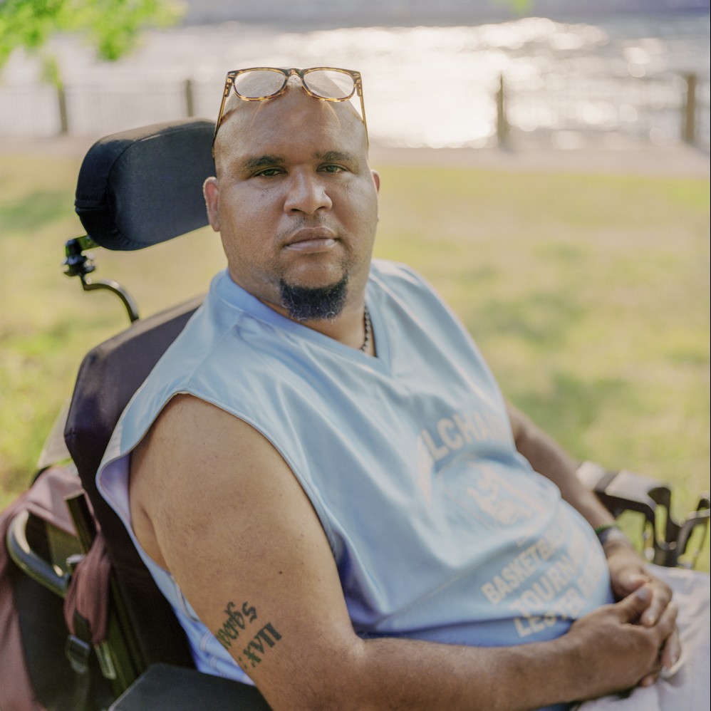 A photograph shows a portrait of poet and director Andres "Jay" Molina shows him sitting in his wheelchair with his hands folded in front of him. He wears a tank top that shows off a tattoo above his elbow, and glasses on top of his head.