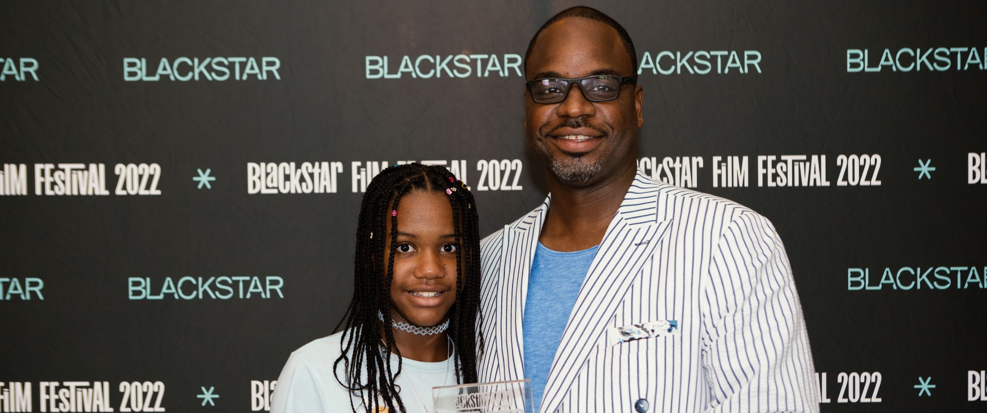 A photo of Bria and Bilal Motley, a Black father-daughter duo, they're standing in-front of a step-and-repeat that says "BlackStar Film Festival 2022." They are holding an award.