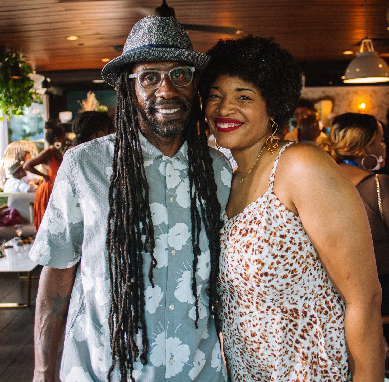 Two people, a Black man with long locs and a Black person with a fro and a dress, are standing together smiling at the camera.