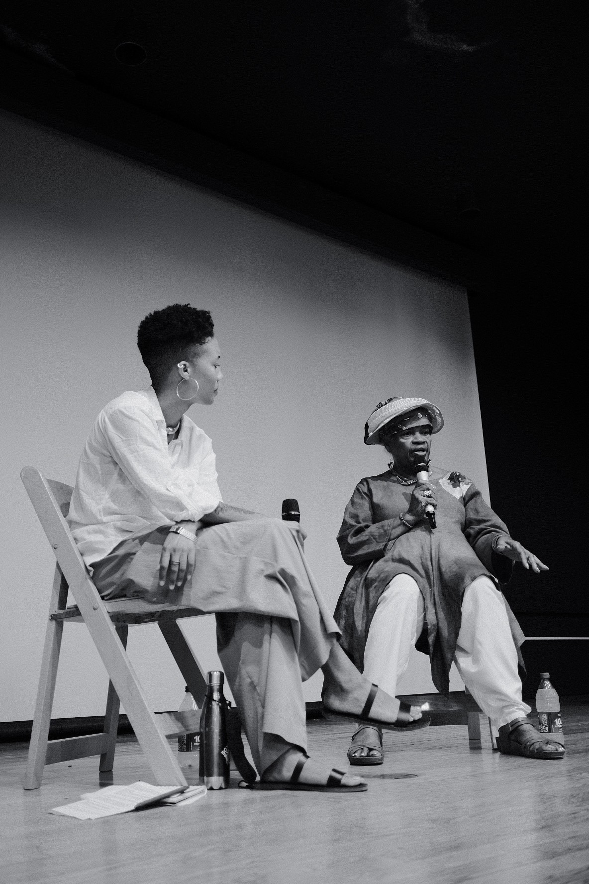 A black-and-white photo of Dessane Lopez Cassell sitting on stage with Dinga McCannon, they are both holding microphones.