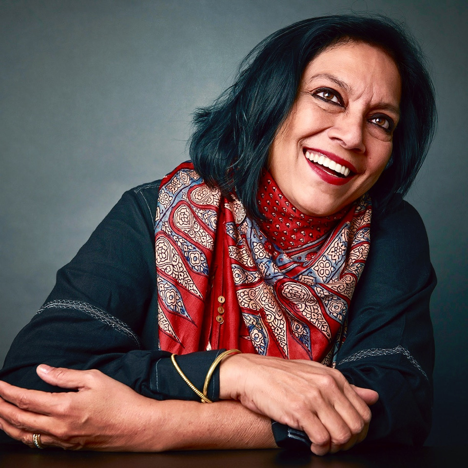 Photo of Mira Nair, she is wearing a black shirt and a red scarf that matches her red lipstick. She has a big smile on her face and looks very happy.