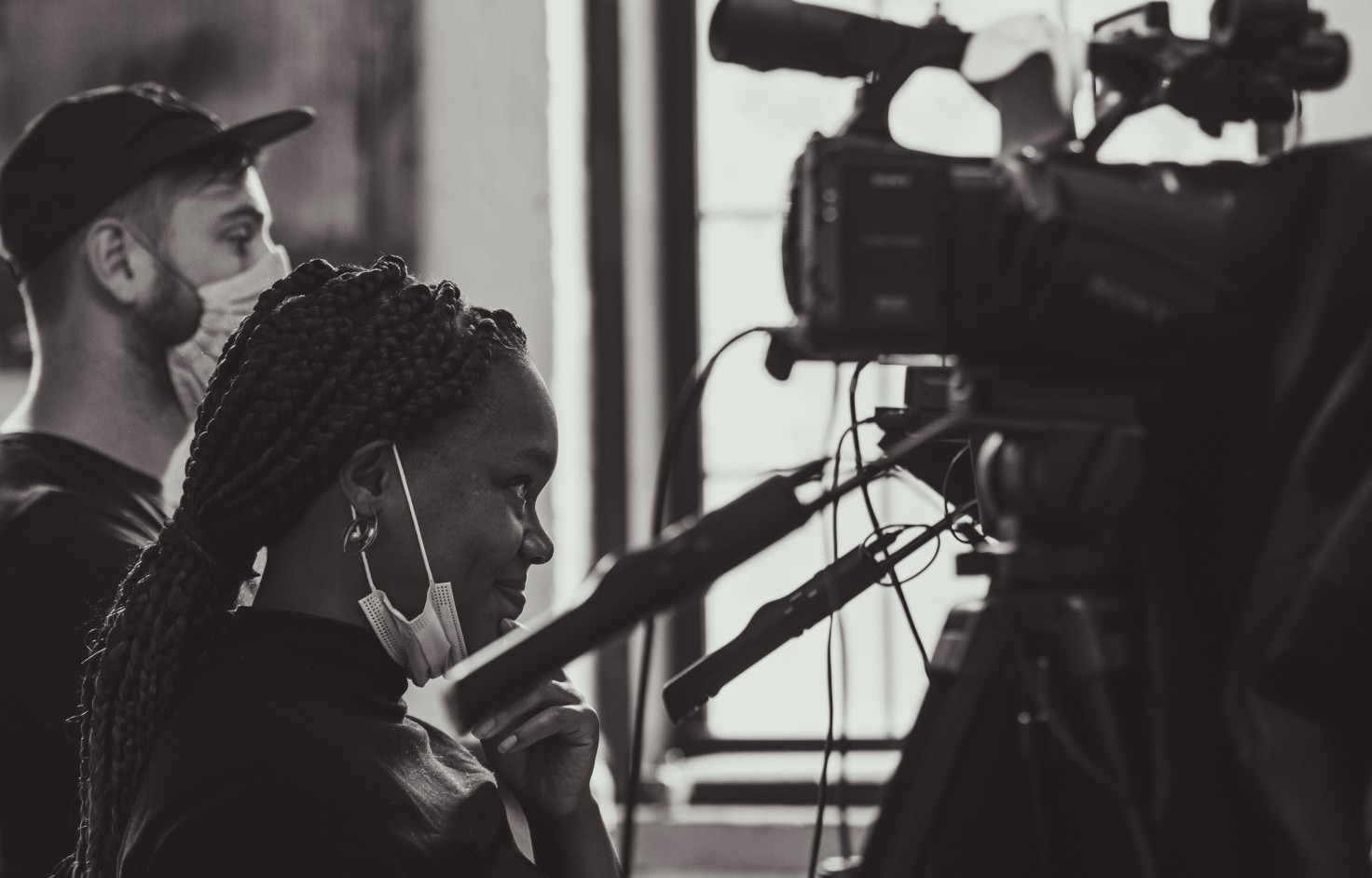 A black-and-white photo of a Black woman controlling a video camera. Her face mask is down and she is smiling.