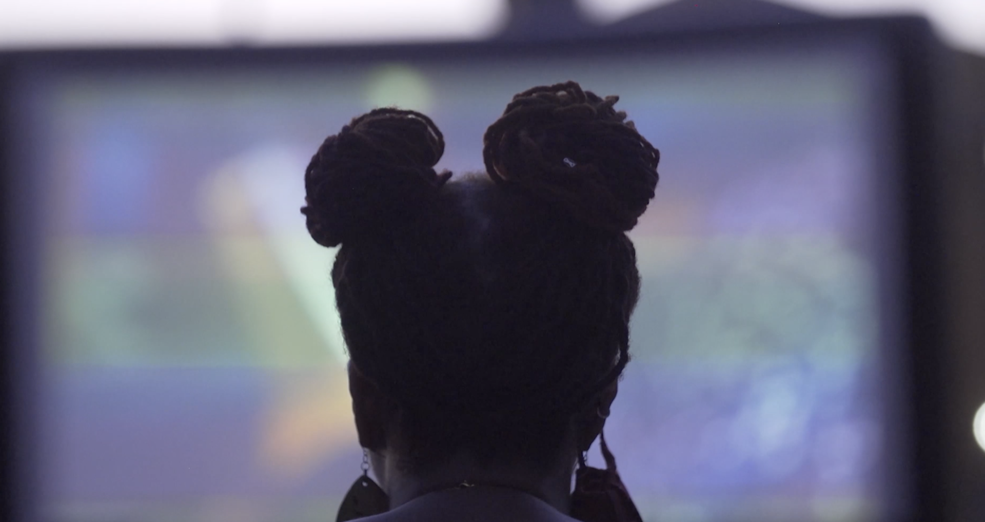 A person with two buns in their hair watches something on a large screen (blurry in the background) as we see them from behind silhouetted.