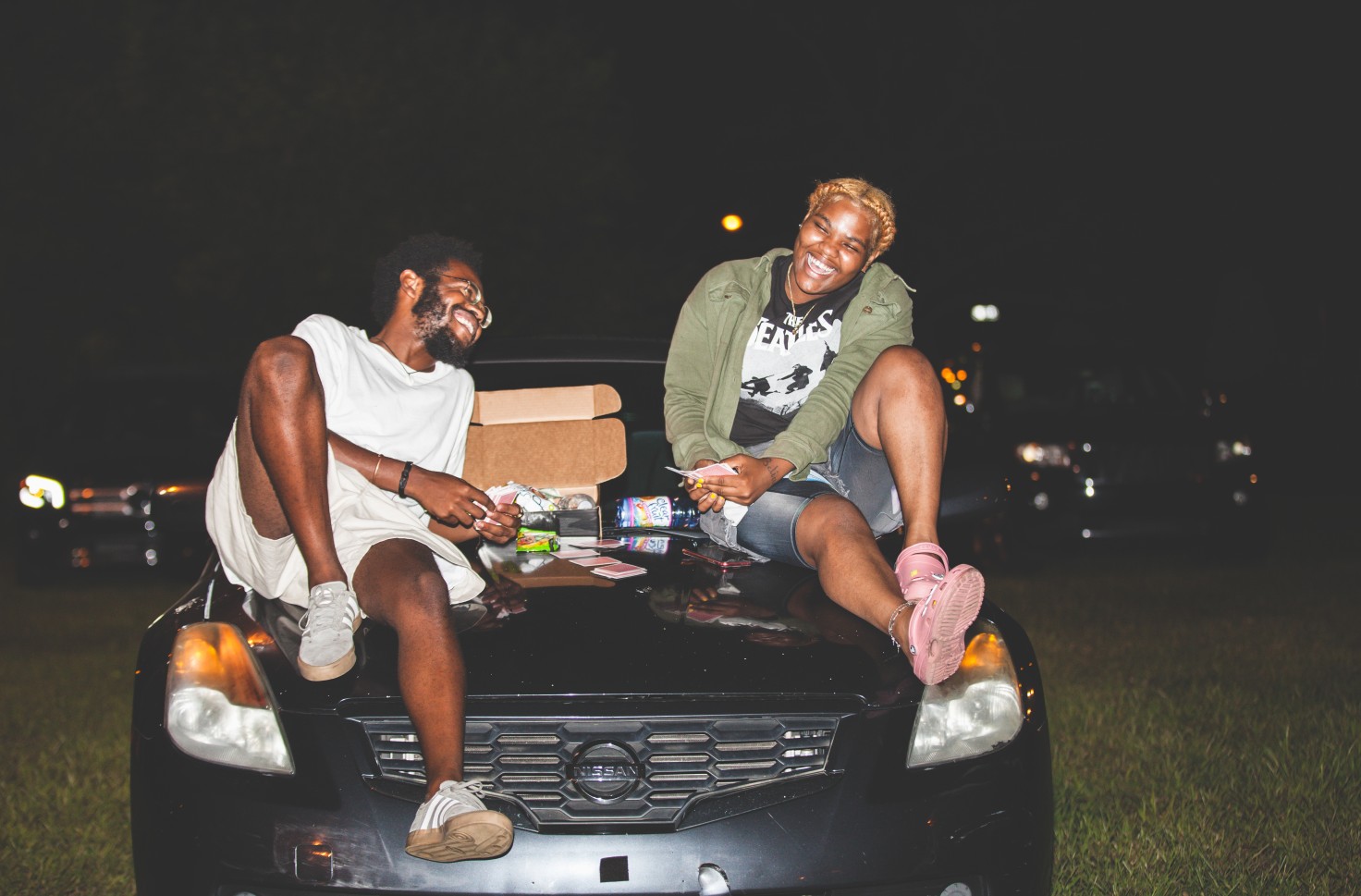 A photo of two Black people sitting on the hood of a car. They have a box full of snacks in between then.