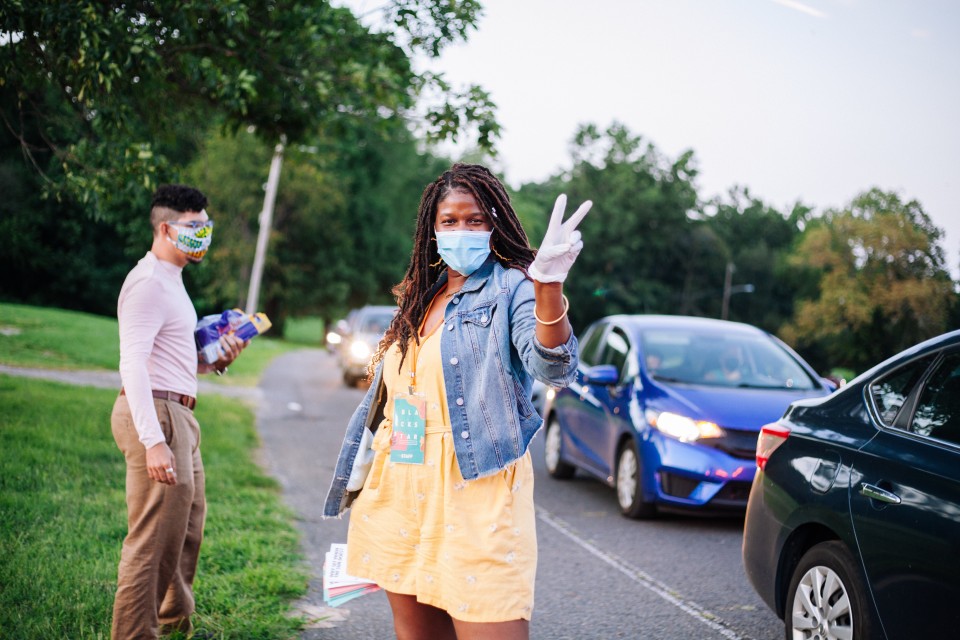 A woman wearing a mask and plastic gloves poses for a picture. Next to her cars are driving down a road presumably towards the drive-in theater.