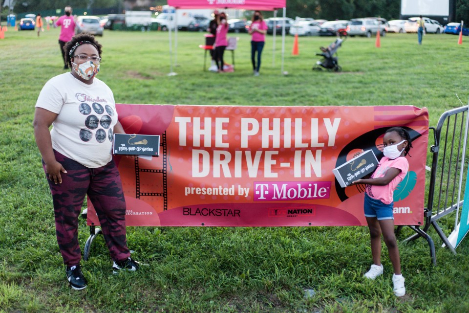 A photo of a Black woman and a young girl posed next to a banner that says "The Philly Drive-In".