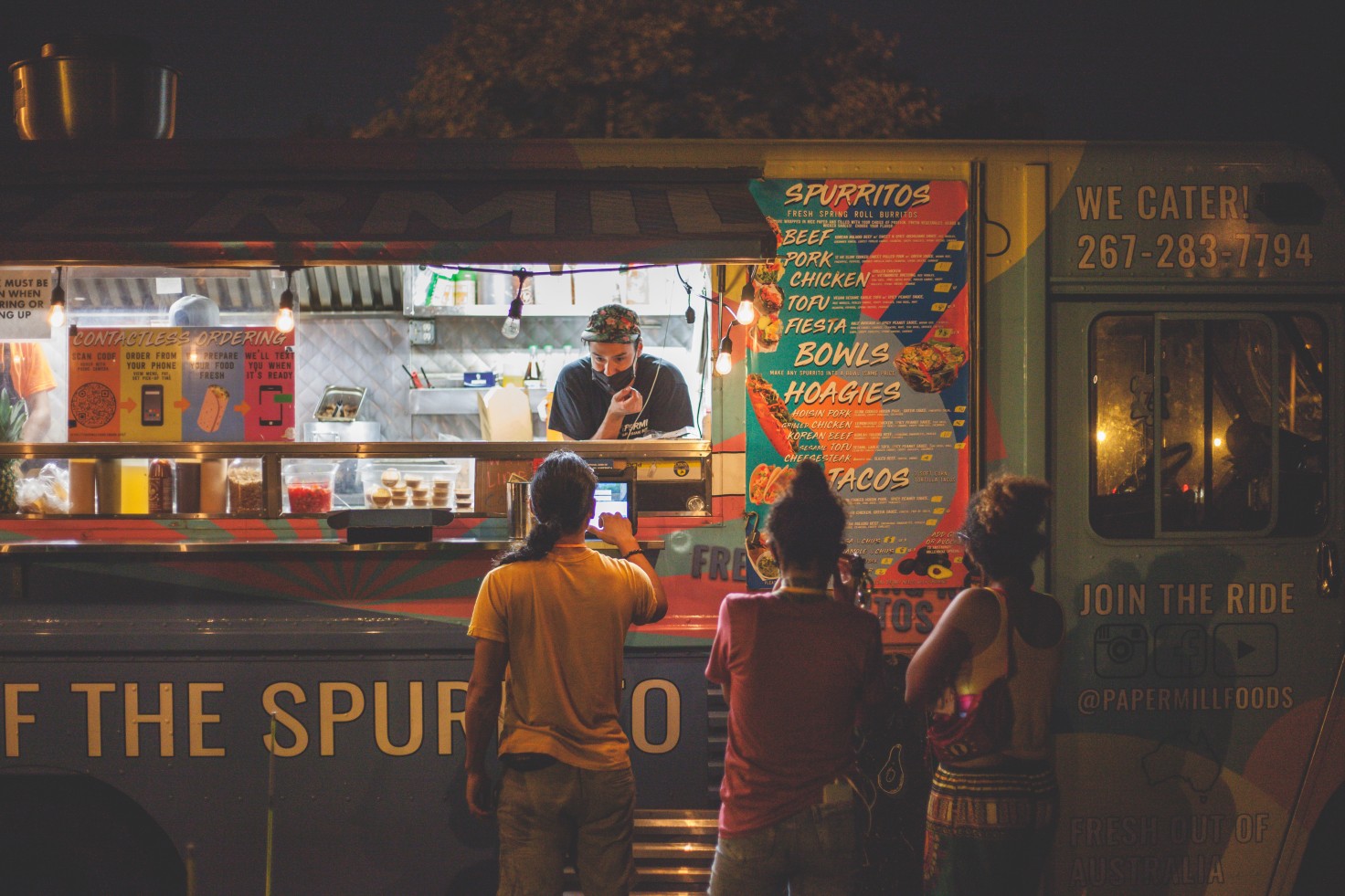 A photo of there people waiting in line at a food truck. The person running the truck is taking the first one's order.