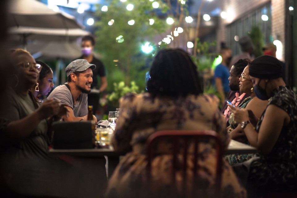 A photo of a group of people sitting at a table outside. There are strands of light around them.