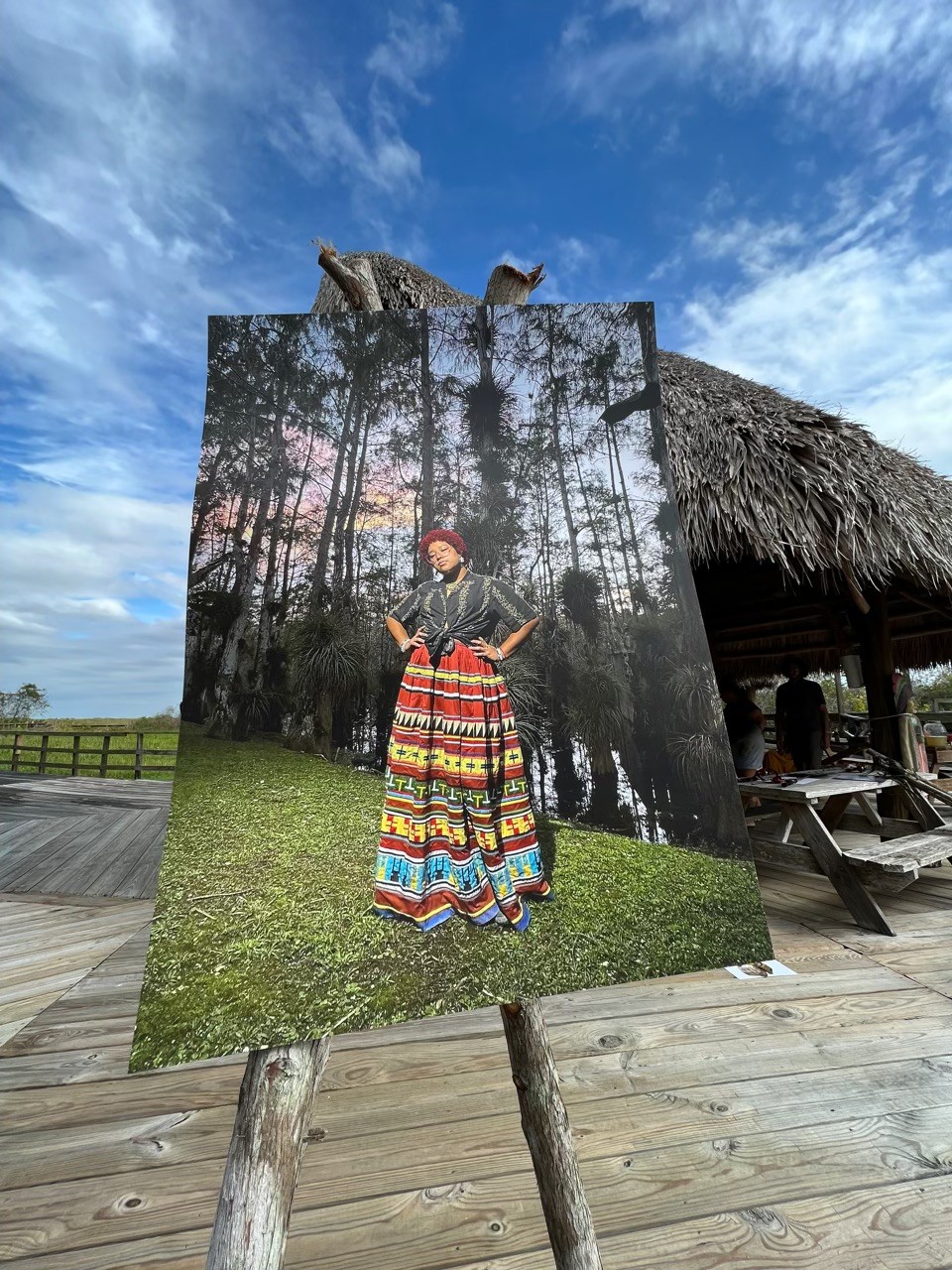 A large photo of a Black and Indigenous person posing on green land with trees behind them is placed on a stand on top of a wooden structure.