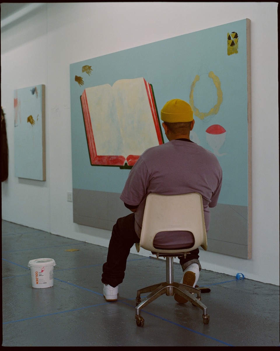 A photo of Kenny Rivero. A black man sitting in a chair with his back to the camera, looking at several large paintings.