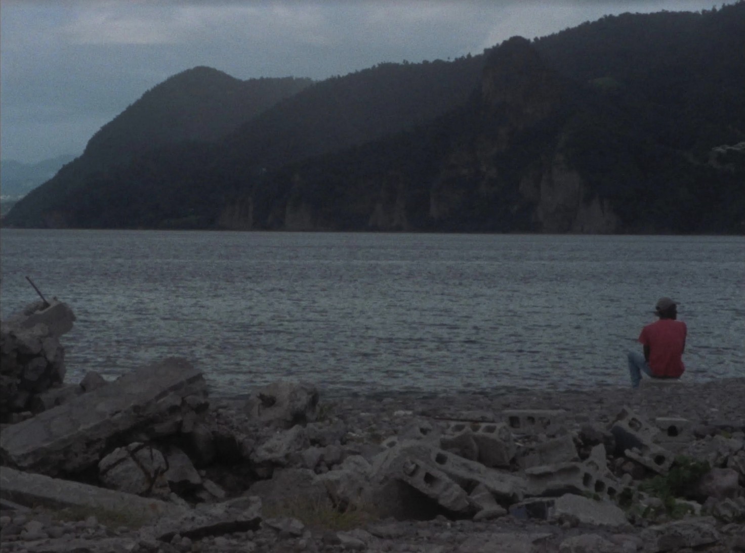 Still from film Chanson for le Nouveau Monde (Song for the New World) directed by Miryam Charles. A landscape of the water, with a figure on the far right in the mid ground.