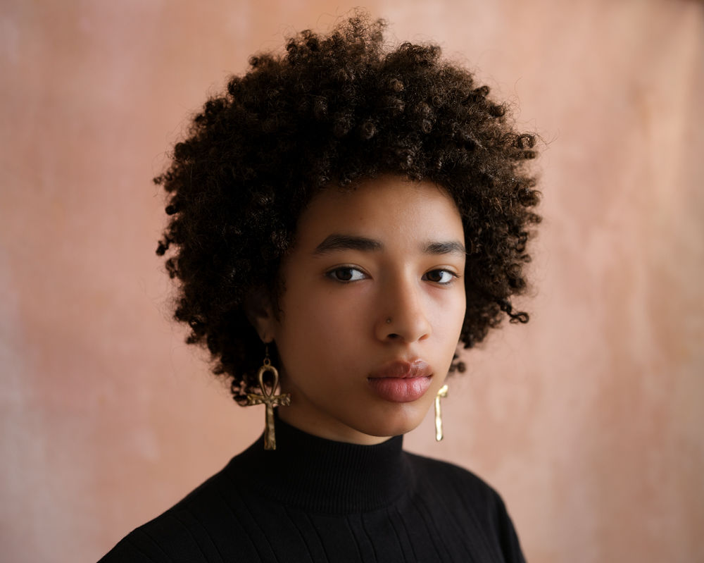 A headshot of Nile. She has an afro and gold earrings. Her expression is straight-faced and focused.