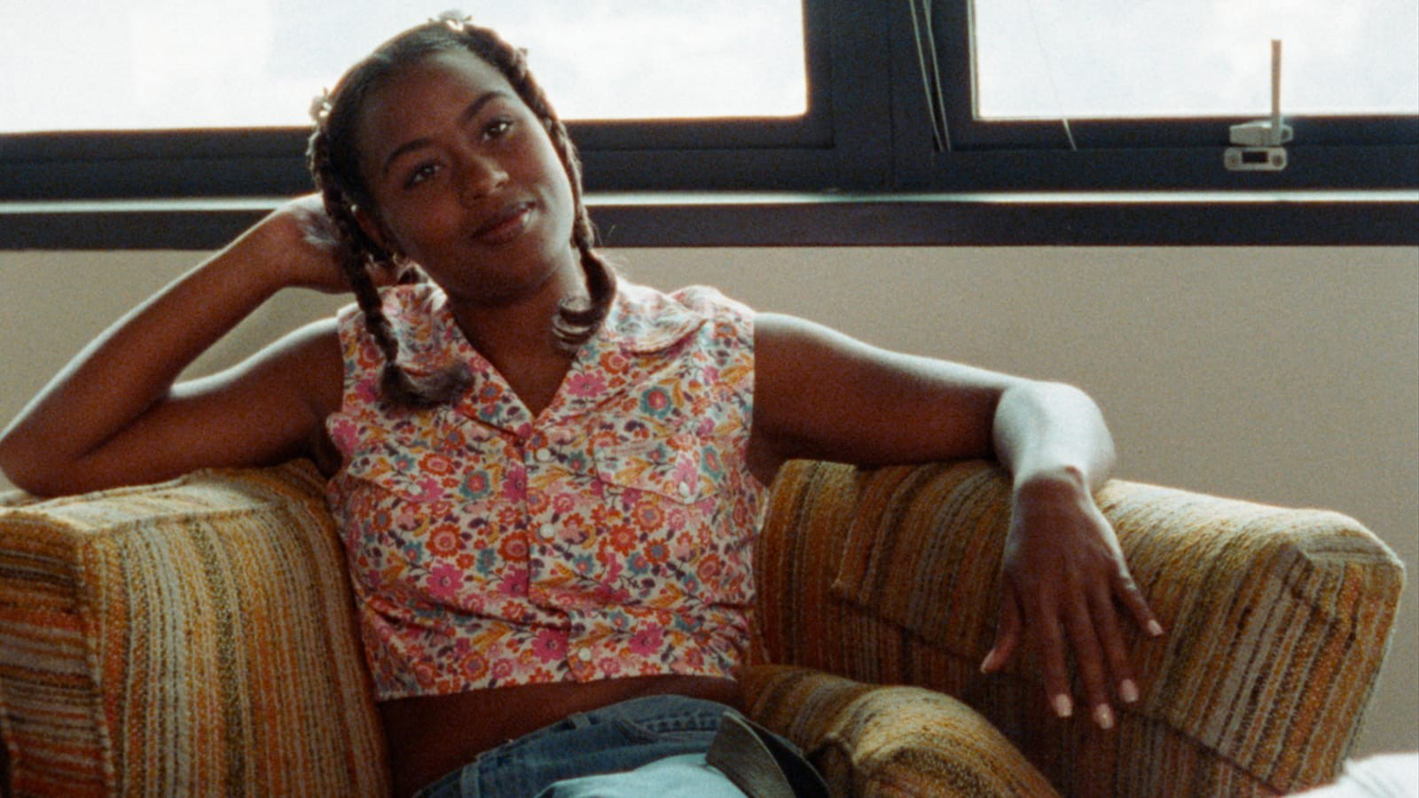 A still from the film Drylongso shows a Black woman reclining on a couch, their head resting on their right hand. They are wearing a flower-patterned blouse and look happy.