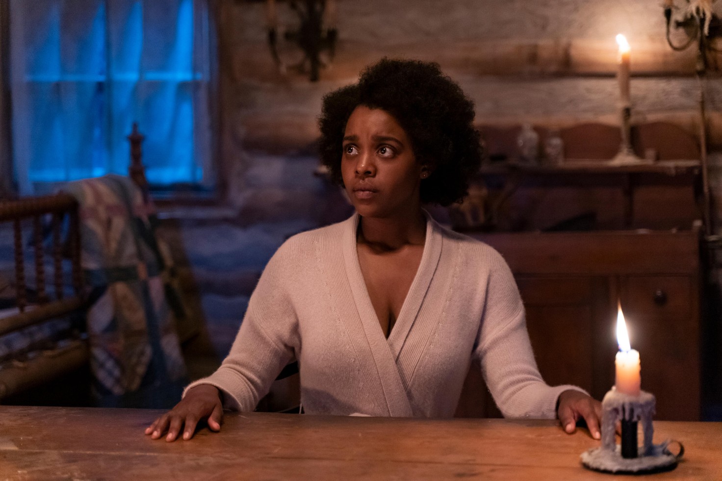 A still from Kindred shows the protagonist — a Black person dressed in modern clothing — sitting pensively at a table which appears to be from a time period in the past.