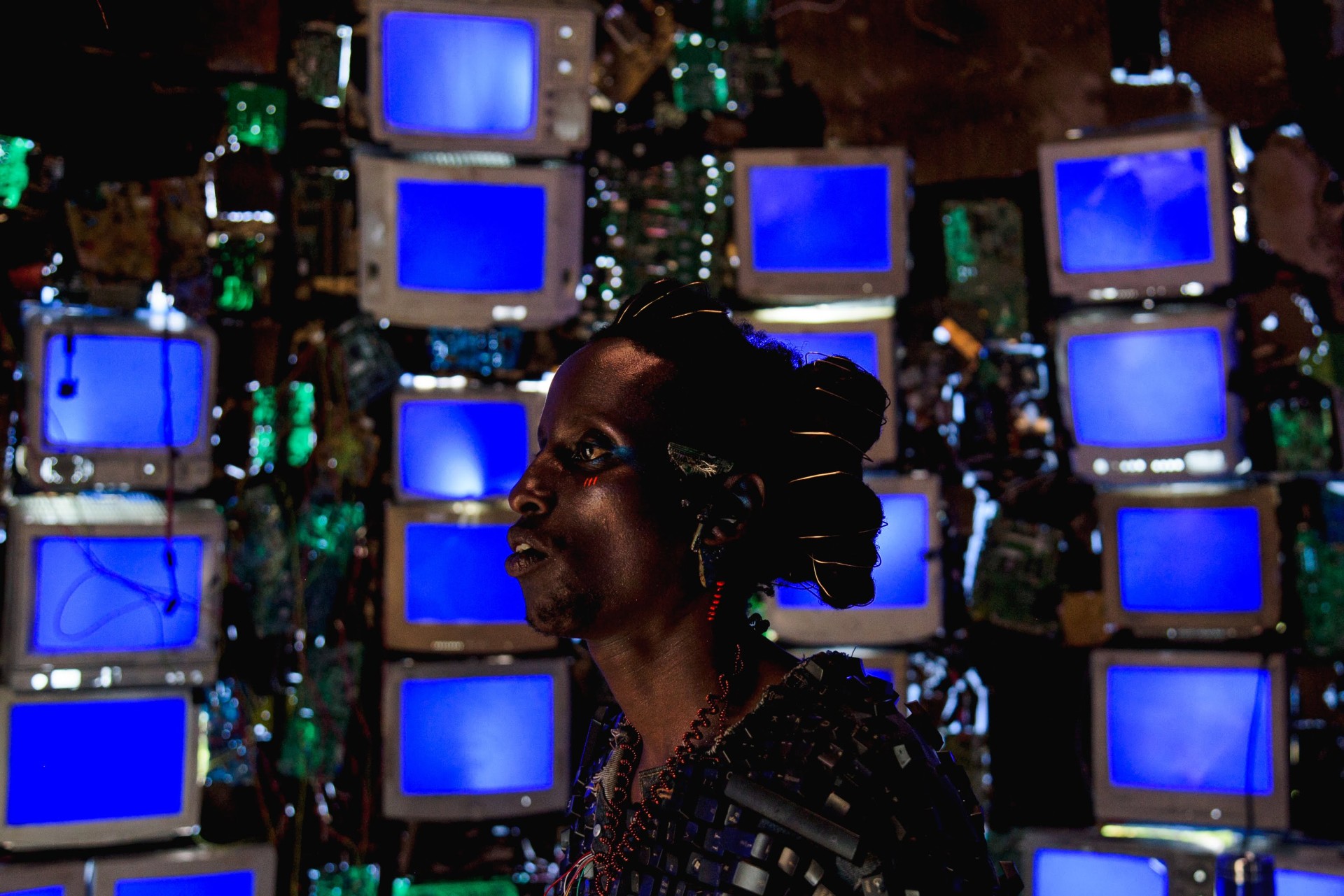 A still from Neptune Frost directed by Saul Williams. A dark skinned person in front of stacks of blue screened computers.
