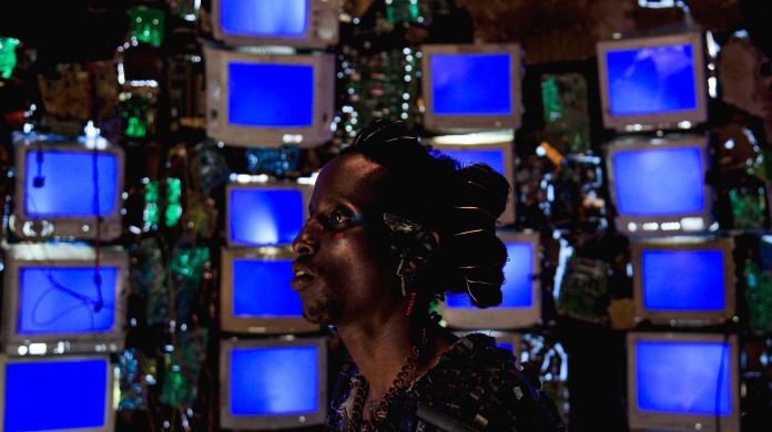 A still from Neptune Frost directed by Saul Williams. A dark skinned person in front of stacks of blue screened computers.