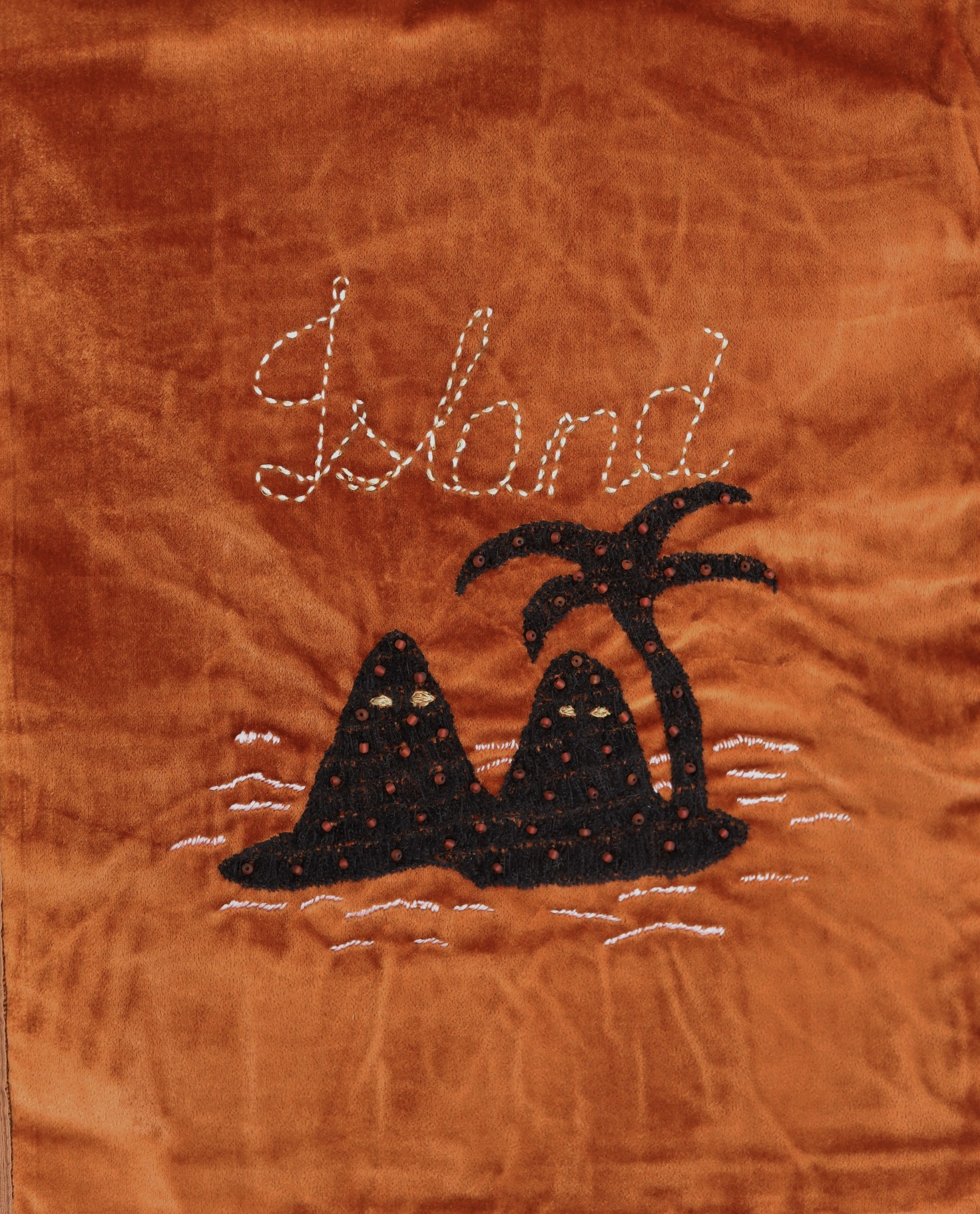 A photo of an artwork by Oneida Rusell. A velvety deep orange fabric with the word Island embroidered in light off-white thread above a fabric silhouette of two mounds and a palm tree.