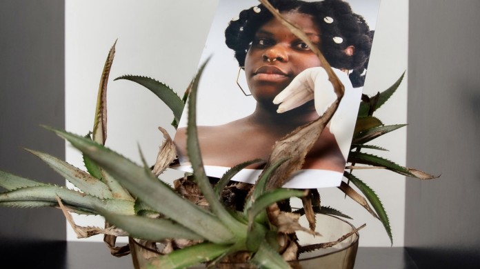 A photo of Sasha Kay's "Sighting Black Girlhood" Exhibition. A printed photo of a young black girl nested in a potted plant.