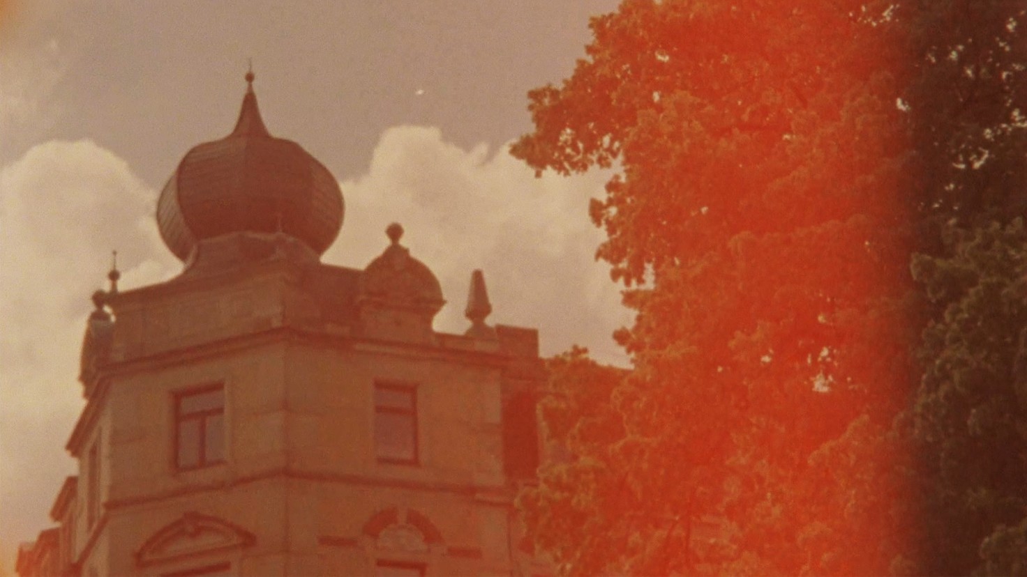 Still from film Drei Atlas (Three Atlas) directed by Miryam Charles. A building with a tree in the foreground, with orange film fading coloring the image