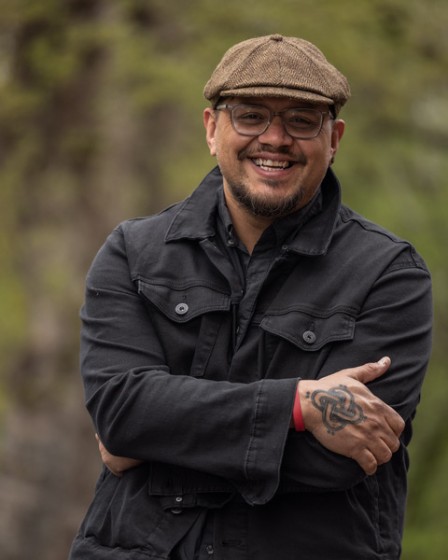 A photo of Sterlin Harjo. He is an Indigenous man. He is standing outside, he is wearing a cap and glasses. He is smiling.