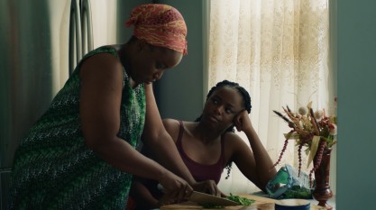 A film still shows two Black people, one older and one younger - perhaps mother and daughter - both indoors. The older person is chopping some vegetables on a cutting board, while the younger person looks up at them, head in hand, in apparent fascination or confusion.