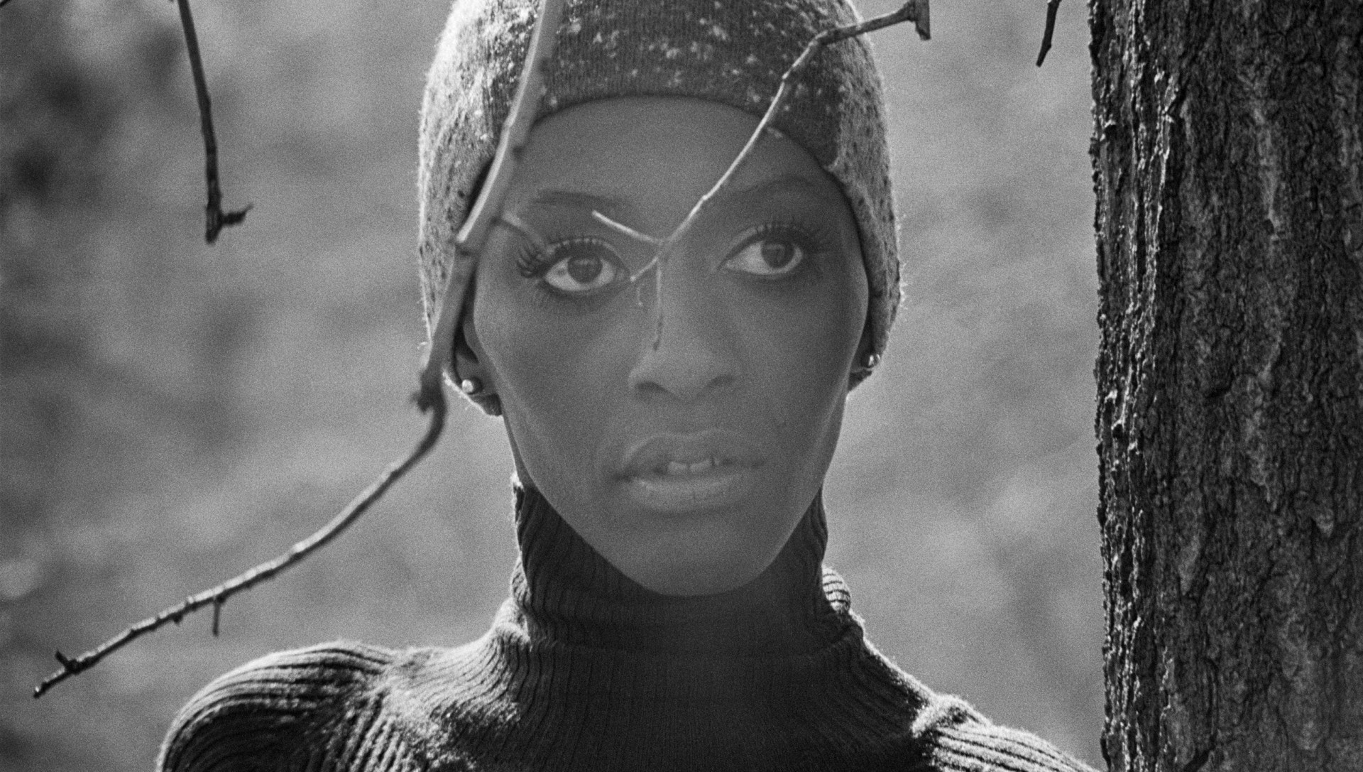 A still photo shows Bethann Hardison, a Black person, in close-up, looking longingly off to the right of the camera, standing behind a tree and wearing a beanie hat.