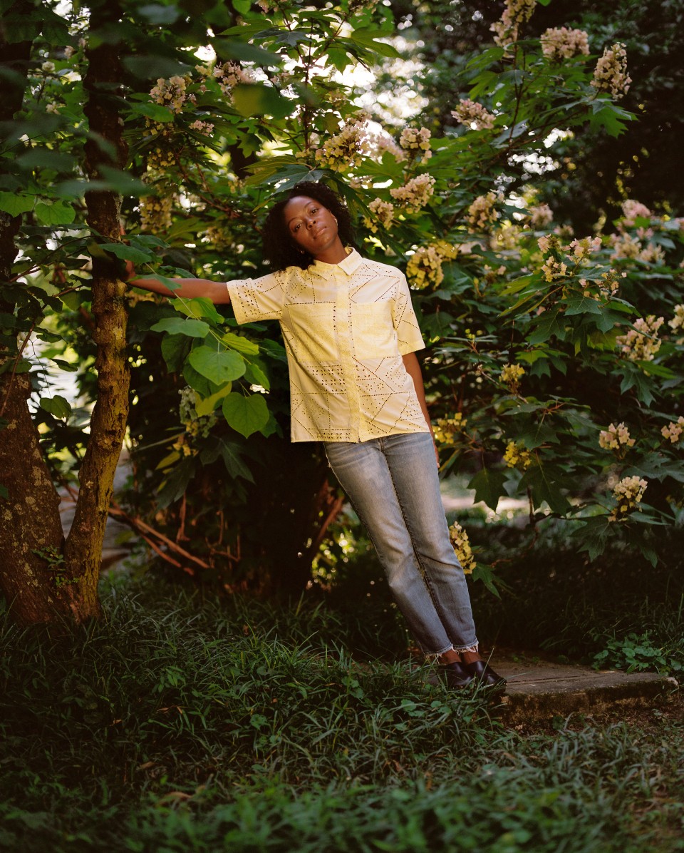 A photo of Danielle Deadwyler who leans against a tree, in a park or forest, wearing a white short sleeve button up and blue jeans.