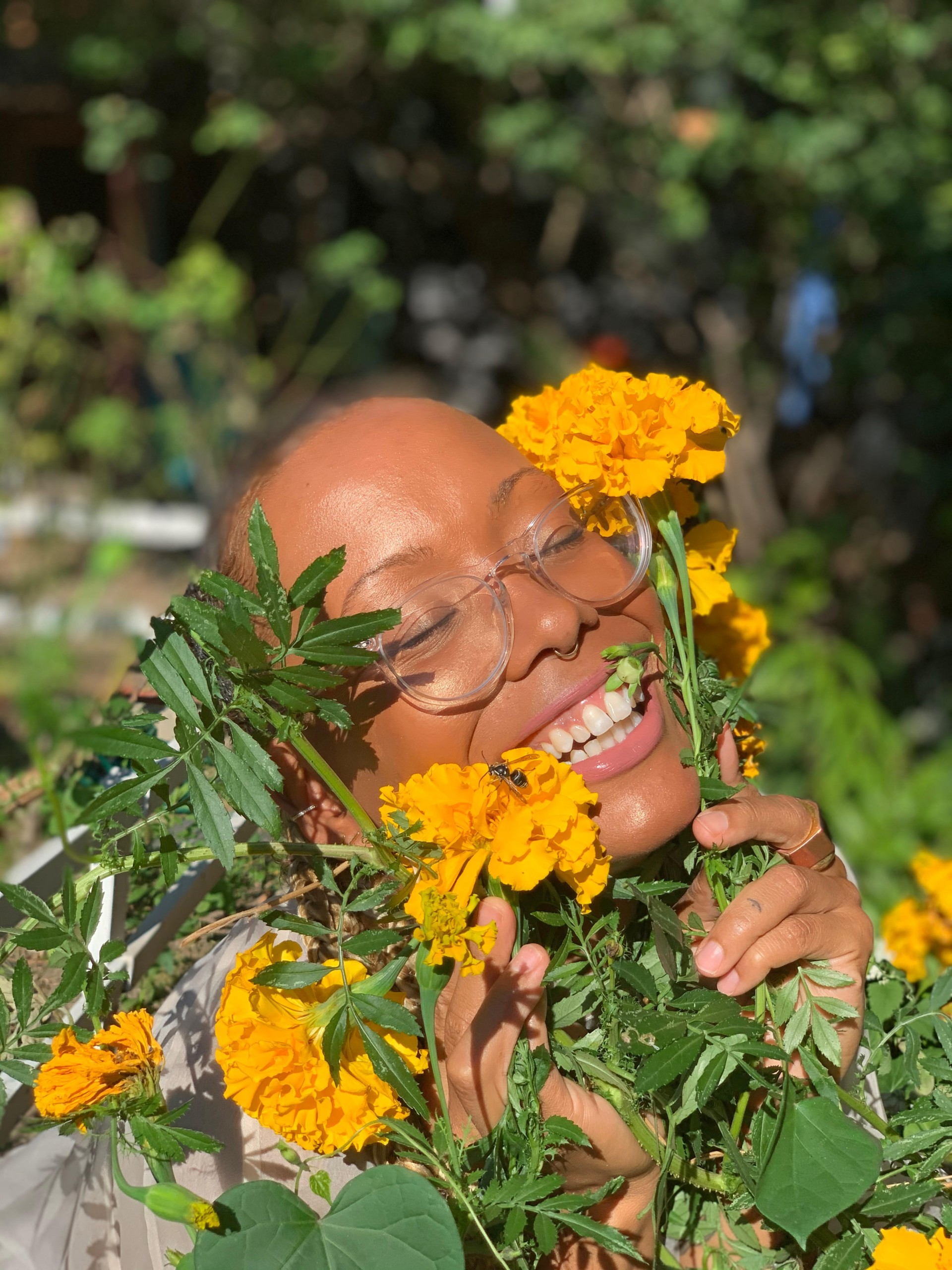 A headshot of J. Wortham. Their face is squished in between marigold flowers. They have glowing brown skin and glasses. They are smiling widely.