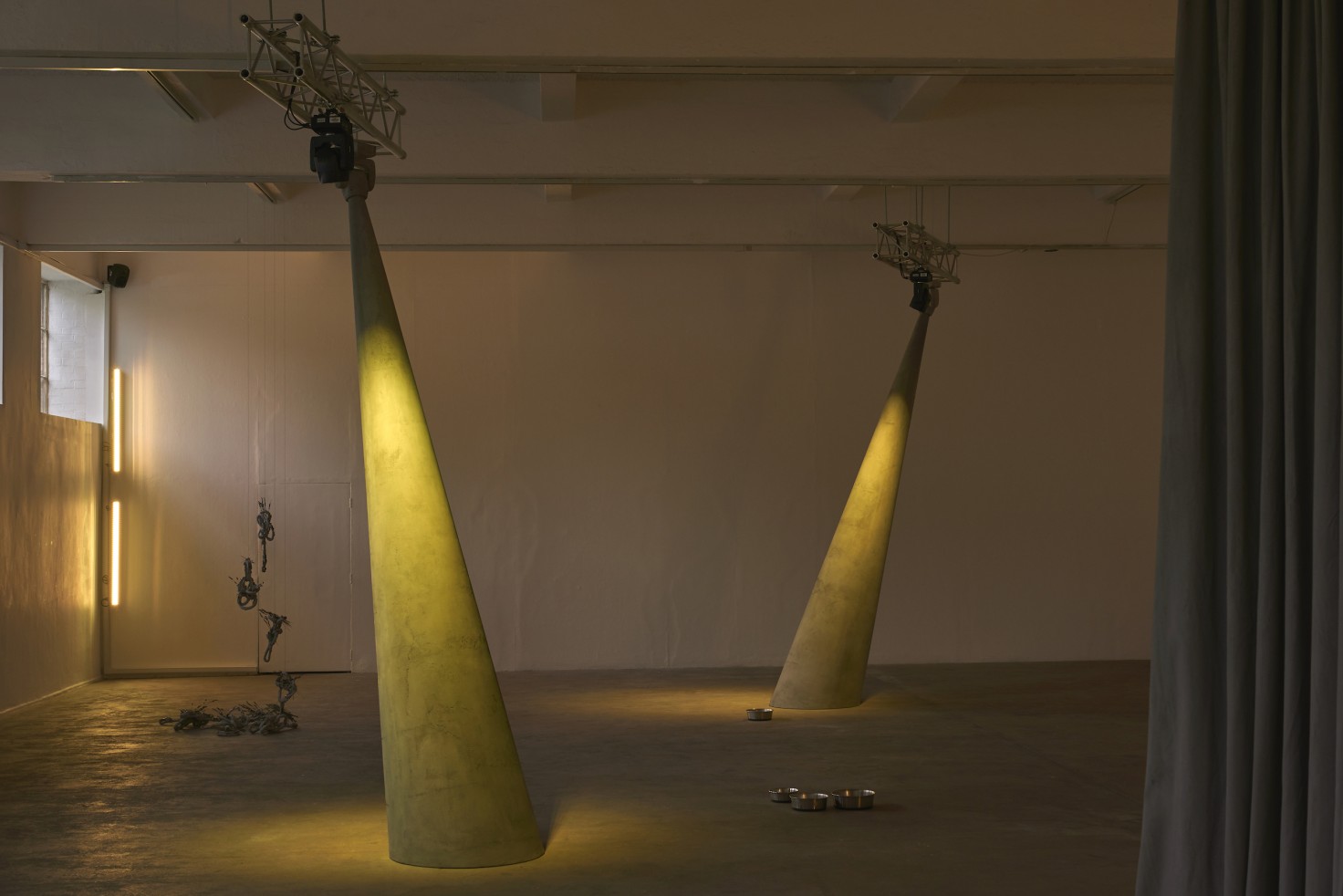 wo concrete columns lit by a warm yellow hue stand in the center of a mostly empty room. On the right hand side a thick curtain occupies around 10% of the frame. On the ceiling, small bits of scaffolding seem to hold up the columns and the two lights that shine down on them. Various aluminum pans lay on the floor and another object hangs in the background on the left hand side.