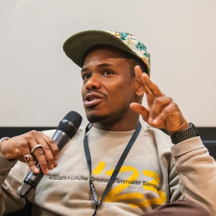 A Black person wearing a baseball hat is speaking into a microphone while wearing a grey sweatshirt.