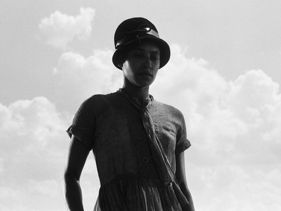 A black and white photo of a woman wearing a hat and a loose cotton dress. Her figure is set against a clear sky with white clouds on the right side of the frame. Her face is partially in shadow and her expression is solemn.