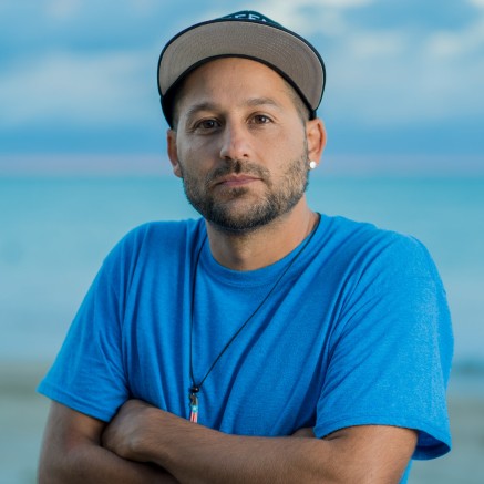A headshot of Eli Jacobs-Fantauzzi, a Puerto Rican man. He is wearing a blue shirt and a baseball cap.