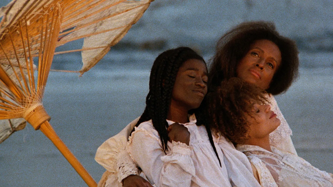 A still from the film Daughters of the Dust by Julie Dash shows three Black women of different ages leaning on each other, all dressed in white, under an umbrella, they look at peace. They seem to be near a body of water as well.