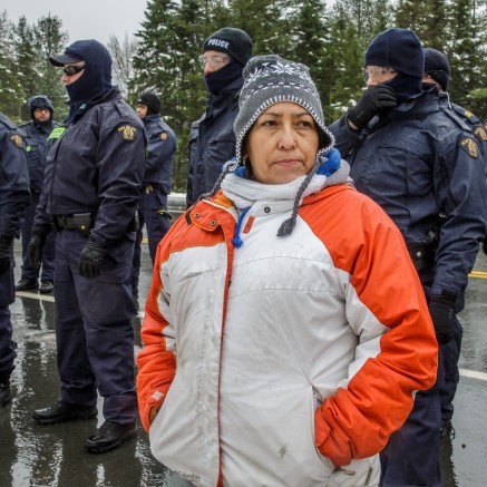 A still from Water Warriors. It shows an Indigenous woman in a winter jacket standing in front of a line of police officers. Her face is contemplative and determined.