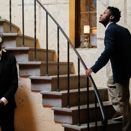 A still from The Freedom to Fall Apart shows two Black men in a mansion-like setting. They are dressed in suits. One is wearing a white mask. The other, who is not wearing one, is looking up a staircase with a curious expression.