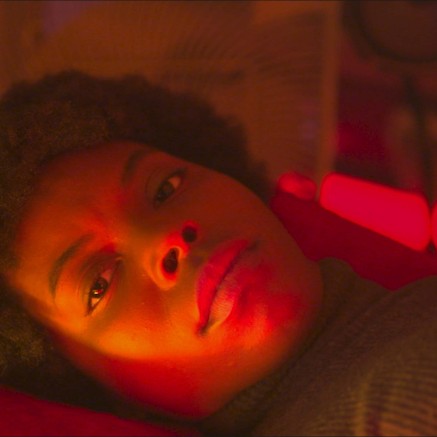 A still from The Love Machine shows a young Black girl laying down. There is a soft red glow on her face.